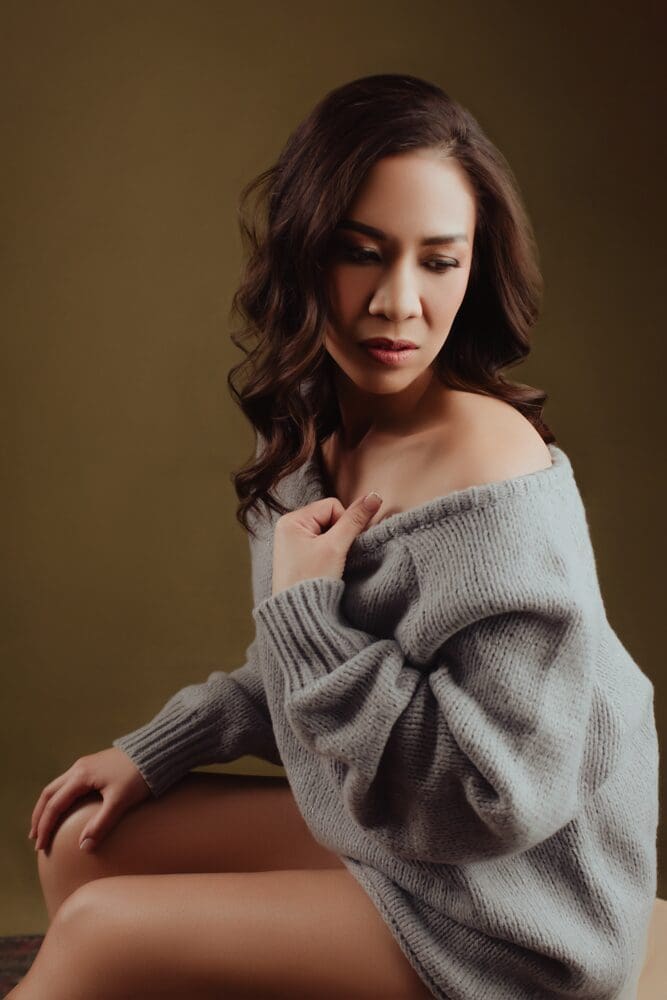 A woman with wavy hair wearing an oversized, off-the-shoulder gray sweater poses against a brown background. She looks downward, expressing a calm, introspective mood.