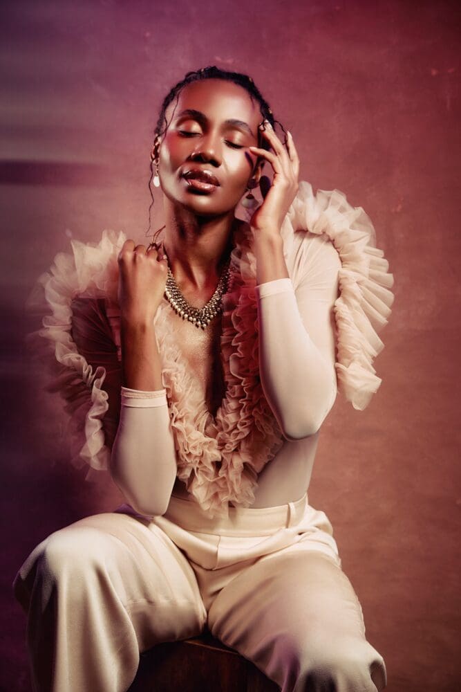 A woman in elegant attire with a ruffled top sits with closed eyes, exuding calm and grace. Soft pink lighting enhances a serene, dreamy ambiance.
