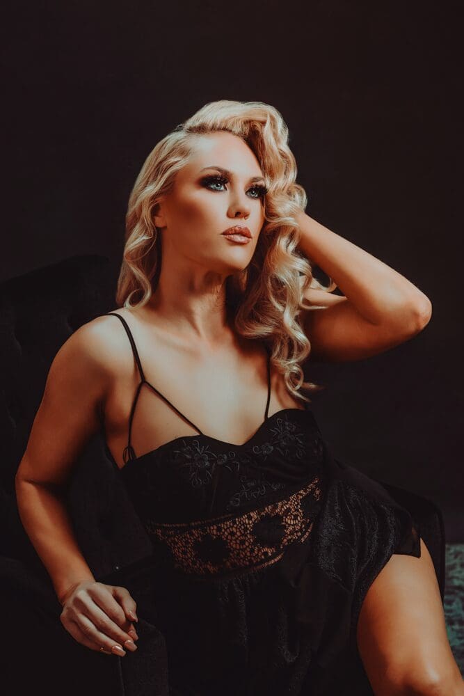 Blonde woman with styled curls sits elegantly, wearing a black lace dress with thin straps. She gazes upwards with a calm, reflective expression.