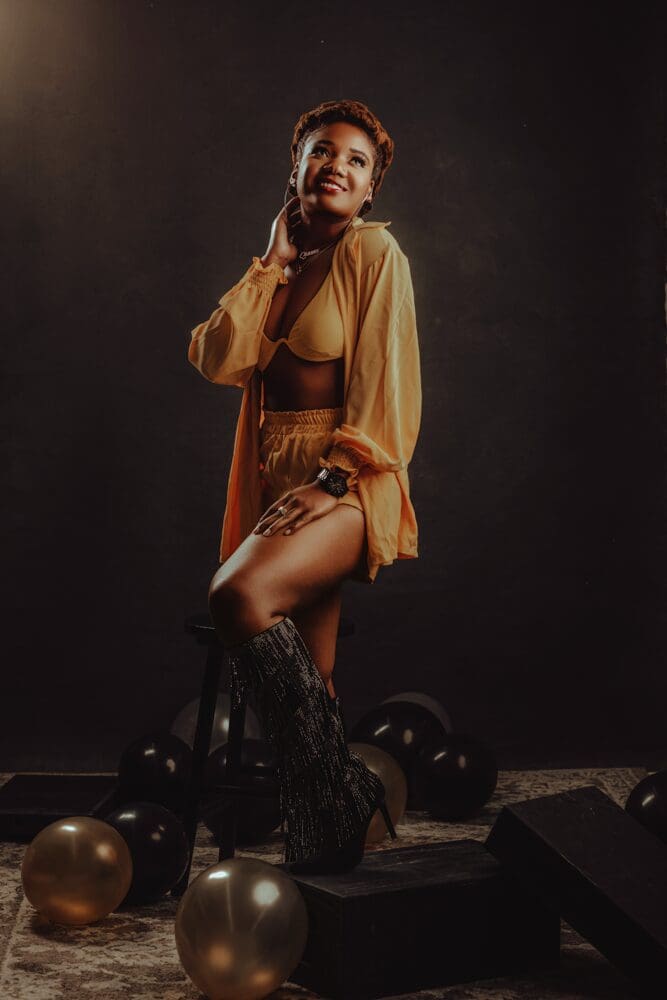 Woman in yellow outfit with layered necklace and hoop earrings, smiling warmly against a dark background. Her pose is confident and relaxed.