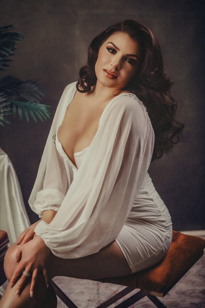 Woman in a white dress with long, voluminous hair sits on a stool, posing gracefully. The background is softly blurred, evoking an elegant, serene mood.