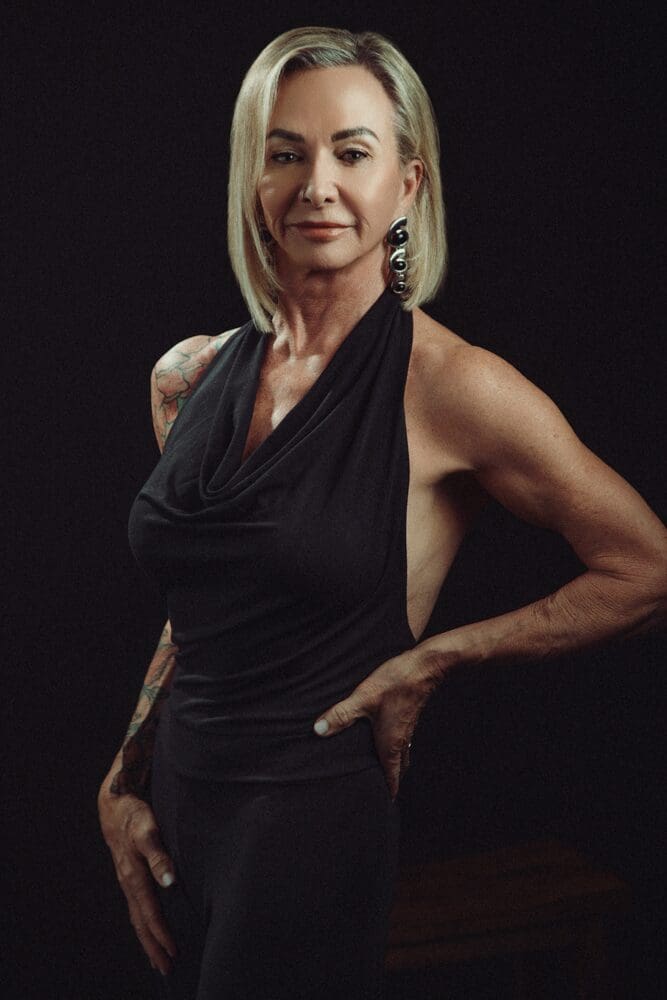 Elegant woman with short blonde hair in a sleeveless black dress, showcasing tattoos and bold earrings, poses confidently against a dark background.