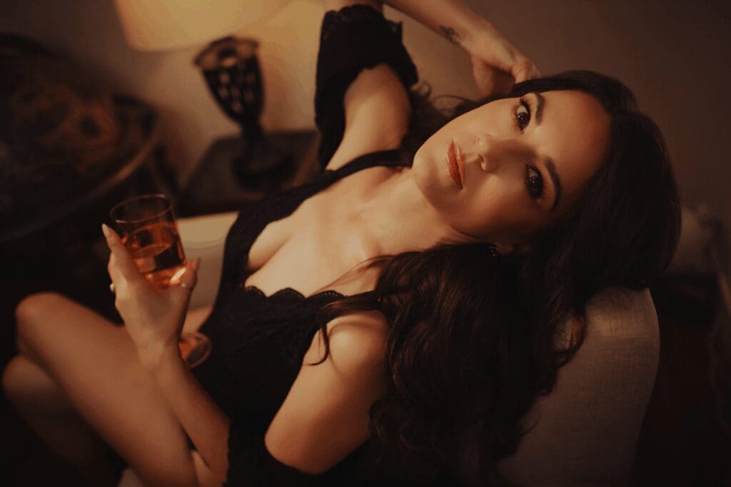 Woman in a black dress reclines on a chair, holding a glass of amber liquid. Soft lighting creates a warm, intimate atmosphere.