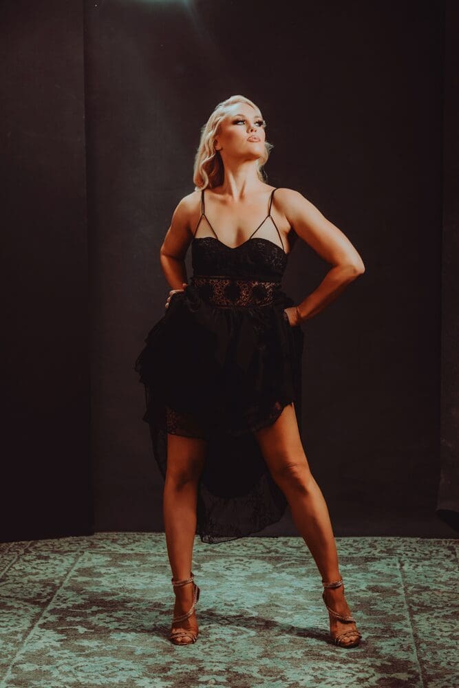 A confident woman stands in a black lace dress with spaghetti straps, hands on hips, on a textured teal carpet. The lighting is dramatic, highlighting her poised expression.