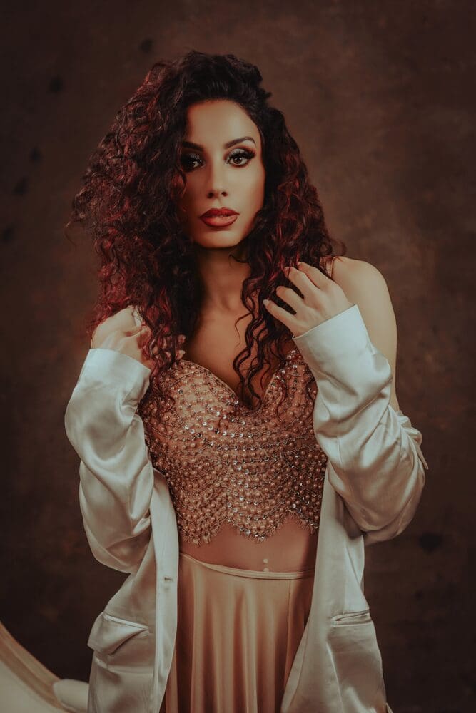 A woman in a lace top and beige coat poses with a gentle smile against a dark background, her hand touching her face. Elegant and serene.