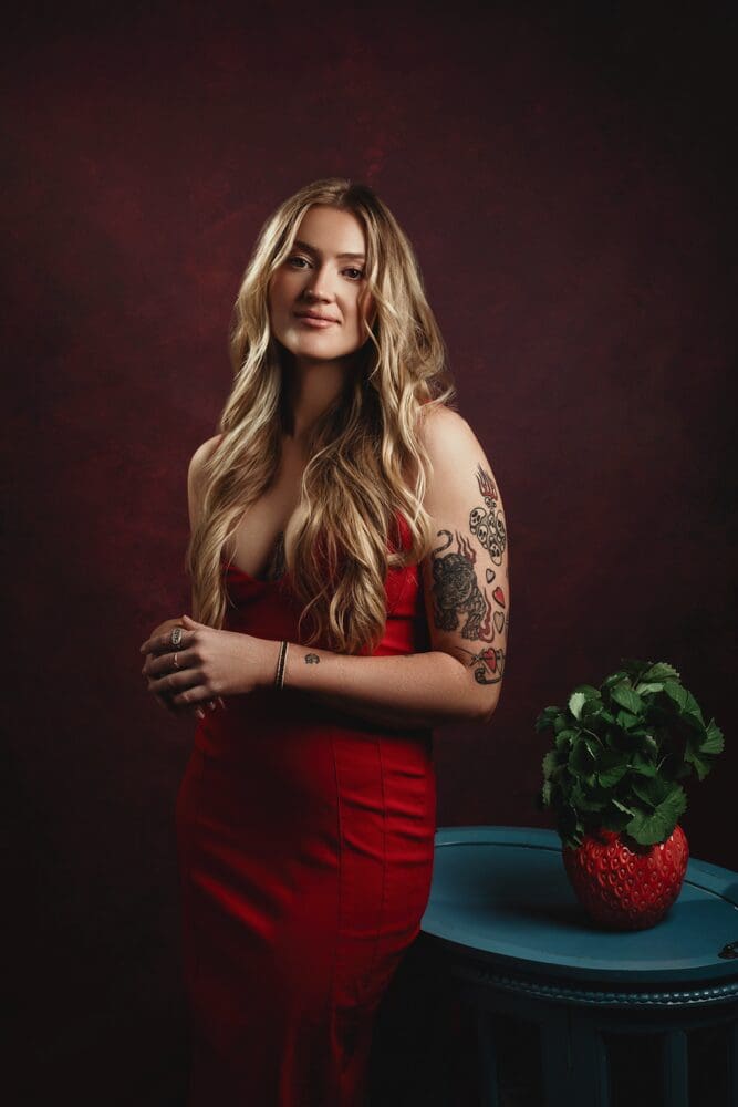 Woman in a red dress with long blonde hair and visible tattoos stands against a dark red background. She appears serene and confident, looking downward.