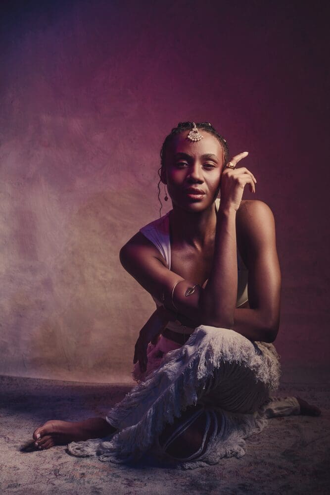 A woman kneels on a textured floor, wearing a fringed skirt and white top. She rests her chin on her hand, with serene expression and dramatic lighting casting a purple hue.