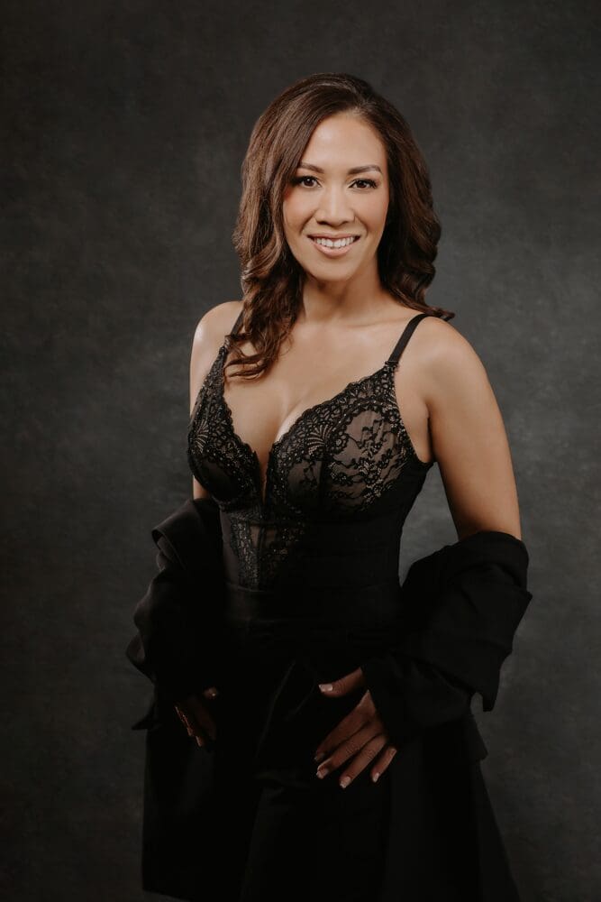 Woman in elegant black lace attire smiles confidently against a dark backdrop. Her wavy hair and relaxed pose convey a sophisticated, poised tone.