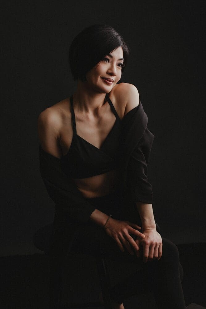 Woman with short black hair in a dark outfit sits on a stool against a dark background, exuding a calm and confident demeanor.