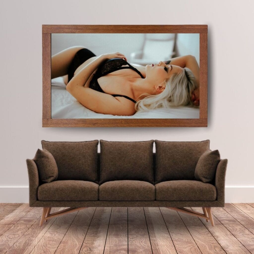 A modern living room with a large framed photo above a brown sofa on a wooden floor. The photo depicts a woman in black attire, lying on a bed.