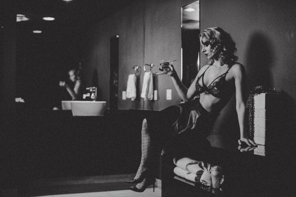 Black and white image of a woman in vintage lingerie, sitting on a bench in a dimly lit bathroom, holding a drink. The scene has a classic, glamorous feel.