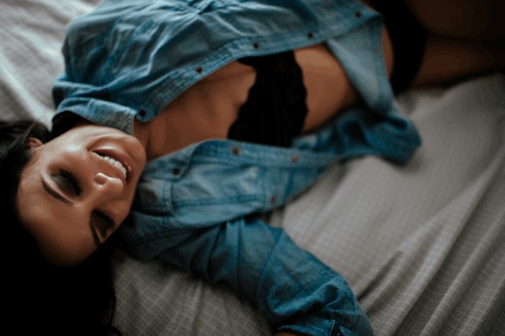 Smiling woman in a relaxed pose on a bed, wearing a denim shirt over a black bra. The mood is intimate and serene, with soft focus and lighting.